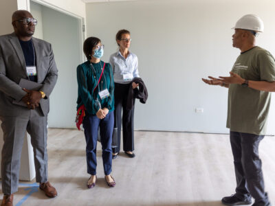 Four people stand in an empty room. The speaker is wearing a hard hat and the others are listening.